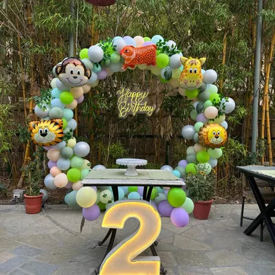 A jungle-themed second birthday setup in an outdoor space. The circular balloon arch is decorated with animal foil balloons, and a large light-up number '2' sits in front for photos.
