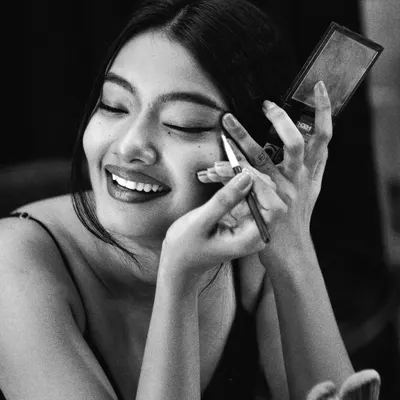 A candid, behind-the-scenes moment captured in timeless black and white. The monochrome treatment emphasizes the joy and emotion of the moment, focusing on the model's smile as she applies her makeup. It tells a story of preparation and passion.