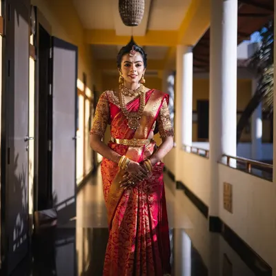 A stunning bride in a classic red Kanchipuram silk saree. The blouse features heavy gold embroidery, and the look is completed with traditional temple jewelry, perfect for a muhurtham ceremony.