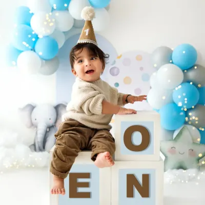 A one-year-old boy sits proudly on blocks that spell "ONE" during his first birthday photoshoot. The blue and white balloon arch adds a festive touch.