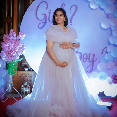 A full-length portrait from a baby shower, featuring a beautiful white tulle gown. The "Girl or Boy?" backdrop and balloon arch create a festive and celebratory atmosphere.