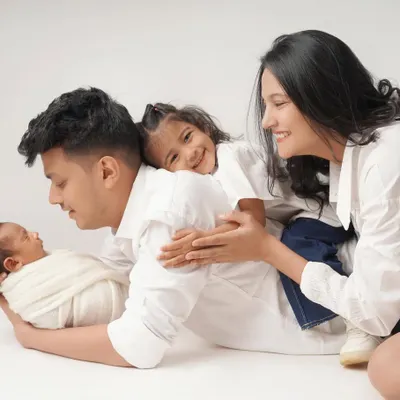 A joyful and playful family portrait with a newborn. The older sister gives her dad a hug while he holds the baby, and mom looks on with a smile. It’s a moment of pure, chaotic, beautiful family life.
