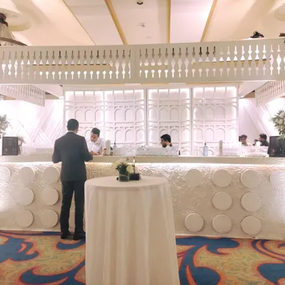 A stunning all-white bar setup for a high-end event. The design features intricate latticework, a textured front panel, and is flanked by large white floral arrangements, creating a sophisticated focal point for serving drinks.