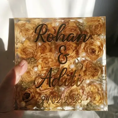 A clear, solid resin block filled with beautifully preserved yellow roses and baby's breath from a wedding varmala. The couple's names are elegantly scripted on the front, making it a timeless decorative piece.
