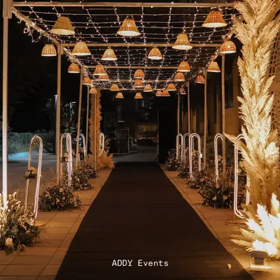 A magical walkway to an evening celebration. This enchanting passage is lit by a canopy of fairy lights and woven basket lamps, with the path flanked by glowing floral arrangements.