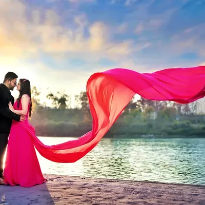 A breathtaking pre-wedding shot by the water, with a vibrant pink fabric catching the wind against the sunset sky. I use elements like flowing dresses to add drama and movement to my photographs.