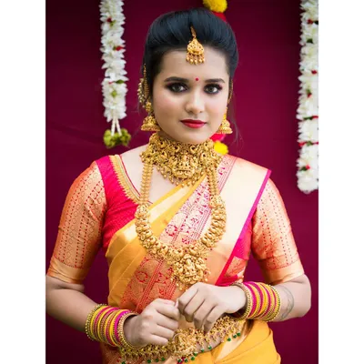 A classic muhurtham look featuring heavy temple jewelry and a bright yellow and red silk saree. The makeup is defined with bold eyes and a red lip to match the traditional attire.