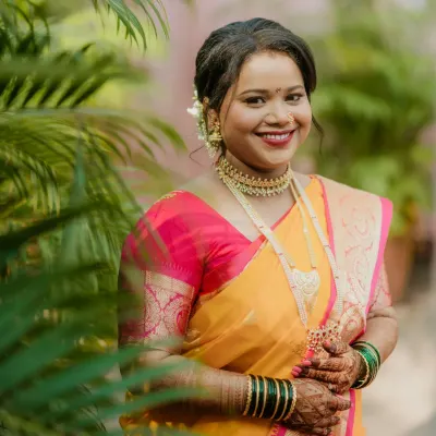 A radiant Maharashtrian bride, captured in natural light. Her look features a warm, glowing base, a classic red lip, and traditional gold jewelry, perfectly complementing her yellow and pink saree.