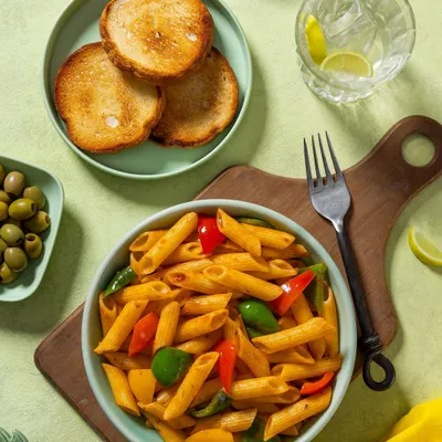 A bowl of penne pasta in a red sauce with bell peppers, shot for Rakesh Masala. The fresh, bright styling with a side of garlic bread makes for an inviting menu image.