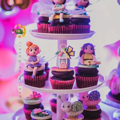 A beautiful display of enchanted forest themed cupcakes. Each cupcake is a work of art, topped with a unique, hand-sculpted figurine like a fairy, a snail, or a butterfly house.