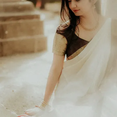 A serene portrait of the bride-to-be, captured in soft, natural light. Her traditional white and gold saree complements the classic stone architecture, creating a moment of quiet elegance.