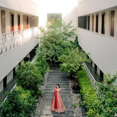 A stunning wide-angle shot of the bride in her red lehenga, framed by the modern architecture and lush greenery of the venue. This portrait showcases both the bride and her beautiful surroundings.