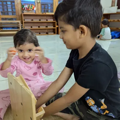 An older brother helps his younger sister with a wooden dressing frame, a practical life activity that develops fine motor skills. This kind of peer teaching happens naturally in our mixed-age group.