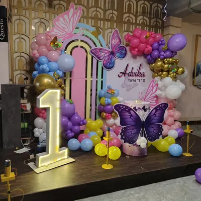 This is a butterfly-themed first birthday stage setup. It features a custom pastel rainbow backdrop, large butterfly cutouts, a light-up number '1', and a colorful balloon garland.
