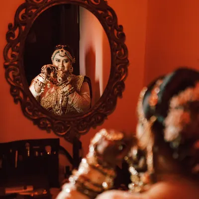 A creative bridal portrait using a mirror to capture the bride's reflection as she gets ready. The warm, reddish lighting adds a dramatic and intimate feel to this getting-ready shot.