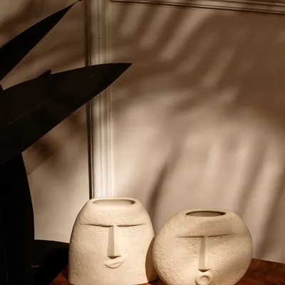 A pair of artistic, face-themed vases styled on a wooden table. The play of natural light and palm leaf shadows creates a warm, serene, and stylish atmosphere.