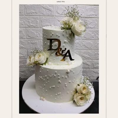This two-tiered white engagement cake is a perfect example of timeless elegance. Adorned with delicate sugar pearls, fresh white roses, and a custom monogram, it’s designed to be a stunning centerpiece for your special day.