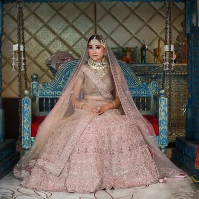 A wide shot of a bride in a magnificent pink lehenga, seated on a traditional blue swing. This composition captures the full grandeur of the bridal ensemble and the ornate setting.