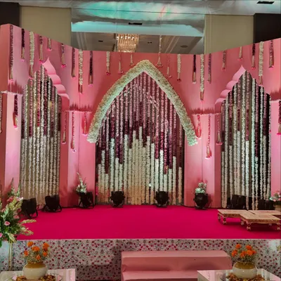 The stage for a 'Bhaat & Tilak' ceremony. The decor uses a blend of traditional elements like floral strings and modern aesthetics with a pink and white color scheme.