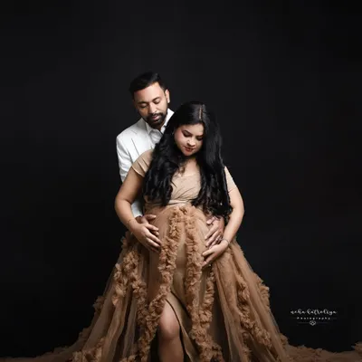 This moody portrait uses a dark backdrop to create a dramatic and intimate feel. The focus is entirely on the couple and the beautiful silhouette of the baby bump, highlighted by the stunning ruffled gown.