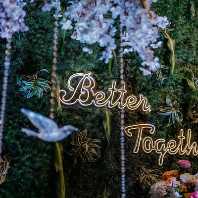 Love is in every detail. This close-up shows our "Better Together" neon sign set against a lush foliage wall, with delicate crystal birds hanging from cherry blossom branches.