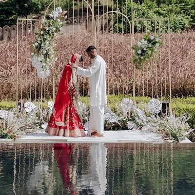 A breathtaking moment at a poolside wedding ceremony. The minimalist golden arch mandap, adorned with white florals, creates a stunning frame for the couple against the natural backdrop.