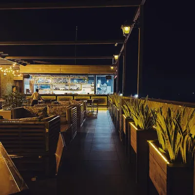 An atmospheric shot of a rooftop cafe at night. The design features an open view into the brightly lit kitchen, slatted wood seating, and a row of illuminated planters creating a pathway.