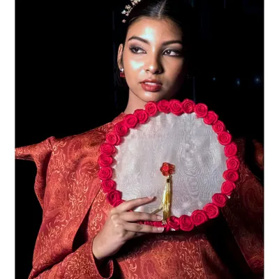 A close-up portrait of a model showcasing an Indo-Western design with a detailed red and gold brocade jacket and an ornate fan. We focus on capturing the texture of the fabric and the model's expression to convey the designer's vision.