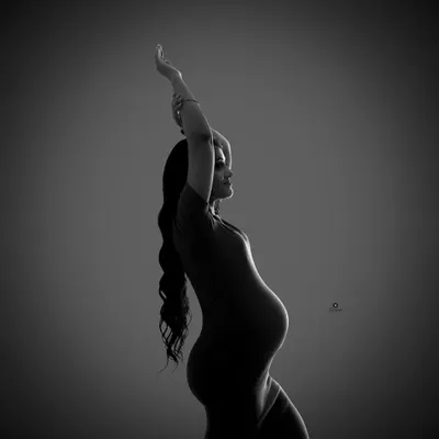 A graceful and artistic silhouette captured in black and white. The raised arm and elegant pose create a beautiful, dance-like shape, highlighting the contours of pregnancy in a unique way.