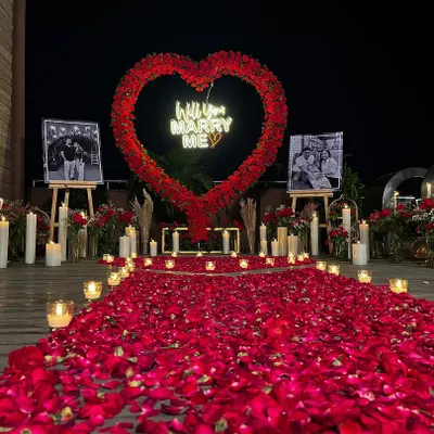A stunning outdoor marriage proposal setup on a terrace. We created this unforgettable moment with a large heart-shaped arch made of red florals, a custom 'Will you MARRY ME' neon sign, and a romantic walkway of rose petals and candles.