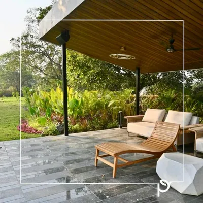 A covered patio overlooking a lush garden we designed. The space features comfortable wooden lounge furniture and is bordered by a dense bed of tropical plants, creating a perfect blend of indoor comfort and outdoor nature.