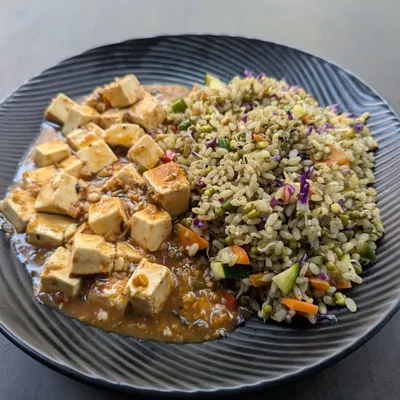 This is a plant-based protein meal featuring tofu in a savory garlic pepper sauce, served alongside pesto-flavored brown rice mixed with vegetables. It's a flavorful and complete vegetarian meal.