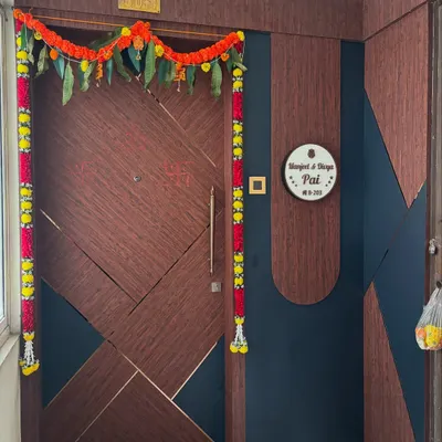 A beautiful main door with a geometric pattern, combining dark wood grain and deep blue panels with fine gold inlay. The entrance is adorned for a housewarming ceremony, showcasing a proud moment for the homeowners.