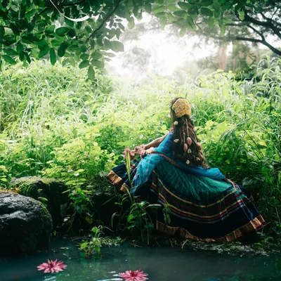 A conceptual portrait capturing the serene and divine essence of Radha, lost in thought by the water in a lush, green forest.