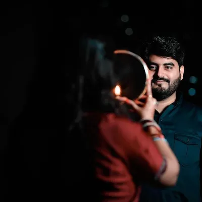 A classic Karwa Chauth moment, where the wife views her husband through the traditional sieve. The low light and the candle's glow create a dramatic, intimate atmosphere, highlighting the husband's loving expression.