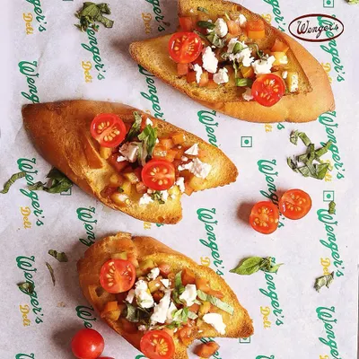 Our Bruschetta is a perfect light snack. This image shows toasted slices of our bread topped with a fresh mix of chopped tomatoes, basil, and feta cheese, served on our iconic deli paper.