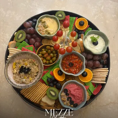 A stunning overhead view of our classic Mezze Platter. It's a colorful mosaic of beetroot hummus, classic hummus, tzatziki, olives, fresh fruits, and assorted crackers, perfect for any social gathering.