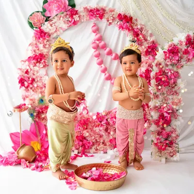 Two adorable little Krishnas. These twin boys are dressed in traditional attire, standing in front of a beautiful pink floral backdrop, making for a divine Janmashtami celebration.