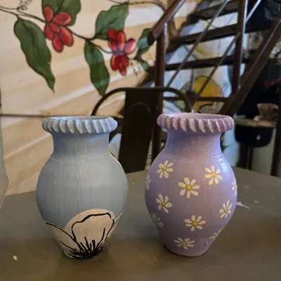 Look at the beautiful details on these finished vases. One features a simple floral outline in a pastel blue, while the other is adorned with hand-painted daisies on a lavender base.