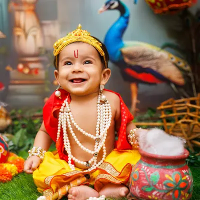 A heart-melting smile from a little Bal Krishna. The setup includes a peacock backdrop, marigold garlands, and traditional props to create an authentic Gokul vibe for this festival costume toddler shoot.