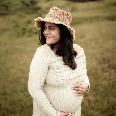 A beautiful, happy portrait of the mom-to-be. Her smile is radiant as she cradles her bump, wearing a straw hat that adds a touch of rustic charm to this outdoor solo shot.