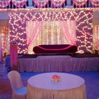 A romantic reception stage featuring a wall of pink flowers illuminated by fairy lights, with a simple red divan sofa, creating a dreamy photo backdrop.