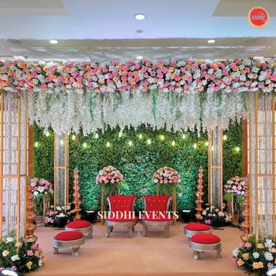 This lush Vidhi Mandap combines a verdant green wall with a canopy of cascading pink and white flowers. The traditional red seating provides a striking contrast, creating a sacred and beautiful space.