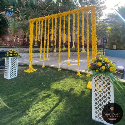 This outdoor Haldi backdrop features a simple frame draped with long strings of yellow marigolds, also known as genda phool. The white latticed pedestals with floral arrangements add a touch of elegance to the sunny, festive setup.