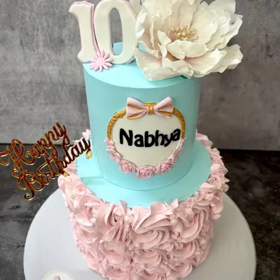 A beautiful two-tier cake for Nabhya's 10th birthday, combining a smooth blue top tier with delicate pink rosettes on the bottom. The large, handmade sugar flower on top adds a touch of elegance.