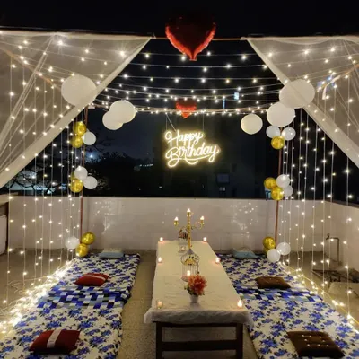 A complete cabana dinner setup on a terrace for a birthday celebration. The structure is draped in sheer fabric and fairy lights, with low seating, candles, and a 'Happy Birthday' neon sign creating a magical dining experience.
