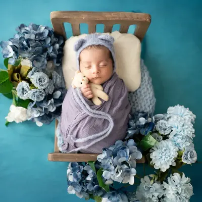 A precious newborn sleeps soundly on a tiny wooden bed, surrounded by a sea of blue flowers. We use creative props and color schemes to design unique newborn portraits.