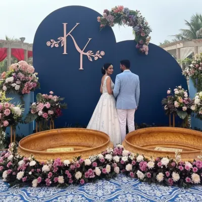 A unique Haldi ceremony setup with a royal blue and pastel theme. The couple is framed by a custom blue backdrop with their initials, surrounded by pink and white floral arrangements and traditional brass urlis.