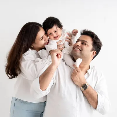A joyful family of three in a classic studio setting. The baby's giggles are infectious as she sits on her dad's shoulders.