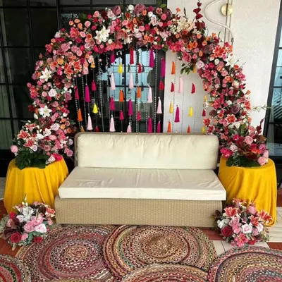 A beautiful seating area I designed for a Mehendi ceremony. The setup is framed by a dense floral arch and a curtain of colorful tassels, with woven rugs adding a bohemian touch.